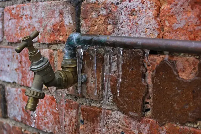 Icicles on an outdoor faucet and pipe along a brick wall in a freeze